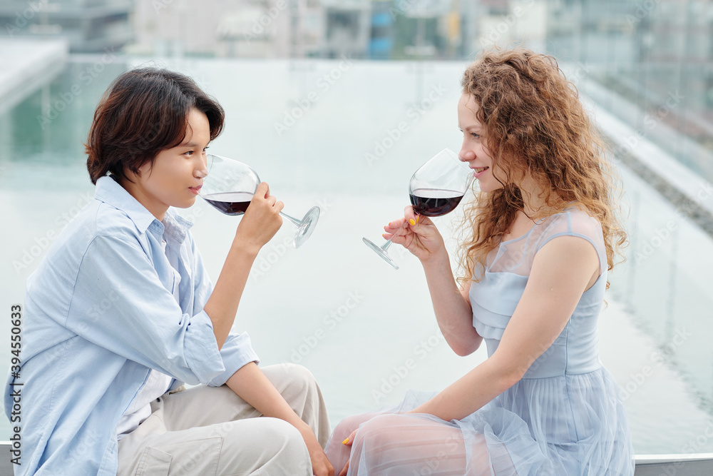 Beautiful young girlfriends drinking red wine and looking at each other when sitting on rooftop