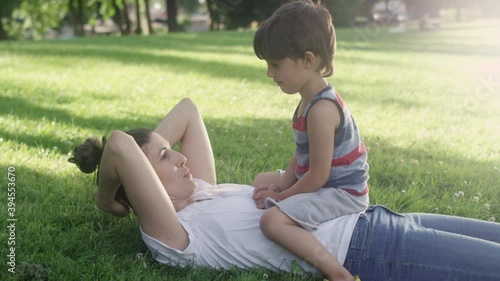 A young boy sits on the stomach of his young mother while laying on the grass at the park