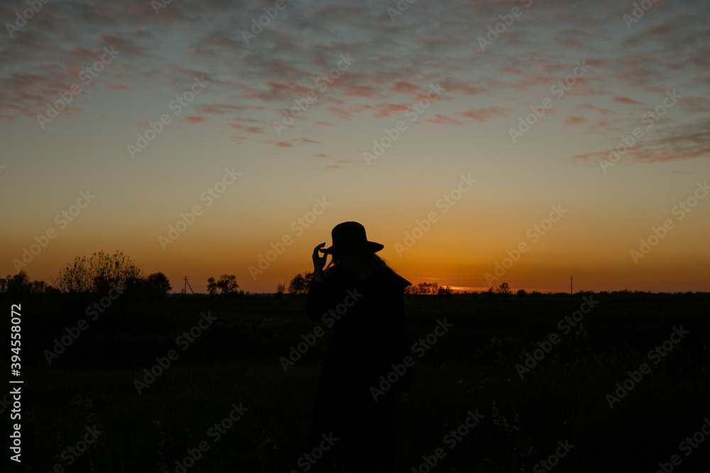 Young sensual woman in black coat and hat  standing in the golden autumn field in the evening on sunset.