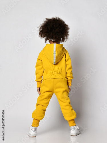 Curly dark-skinned kid girl in warm blue sports jumpsuit with hood and sneakers stands back to camera over white wall background, rear view. Trendy children fashion, stylish outfit