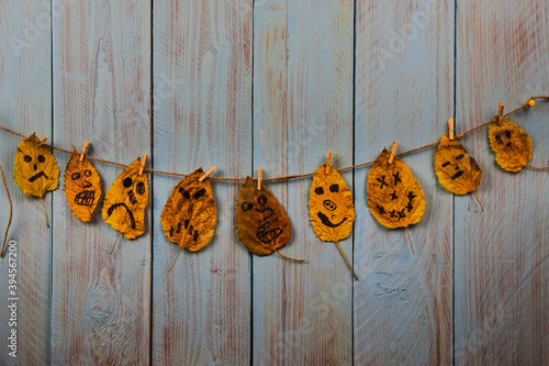 Funny smiley emoticon face drawn on golden autumn leaves hanging on the rope against blue rustic wooden background. Copyspace
