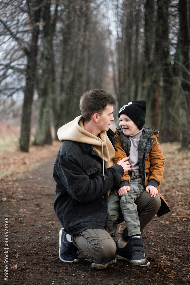 father and son are walking in the park, sitting in the middle of the alley, single father, cold spring, autumn, winter, against the background of large trees, have fun smiling, boy sits on his knees