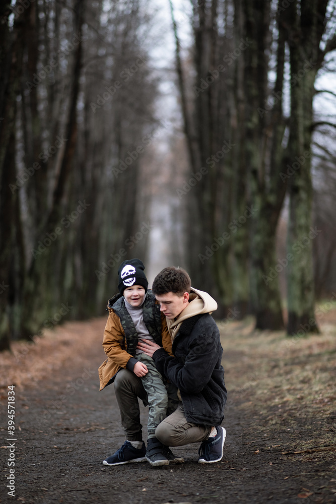 father and son are walking in the park, sitting in the middle of the alley, single father, cold spring, autumn, winter, against the background of large trees, have fun smiling, boy sits on his knees father and son are walking in the park, sitting in the middle of the alley, single father, cold spring, autumn, winter, against the background of large trees, have fun smiling, boy sits on his knees