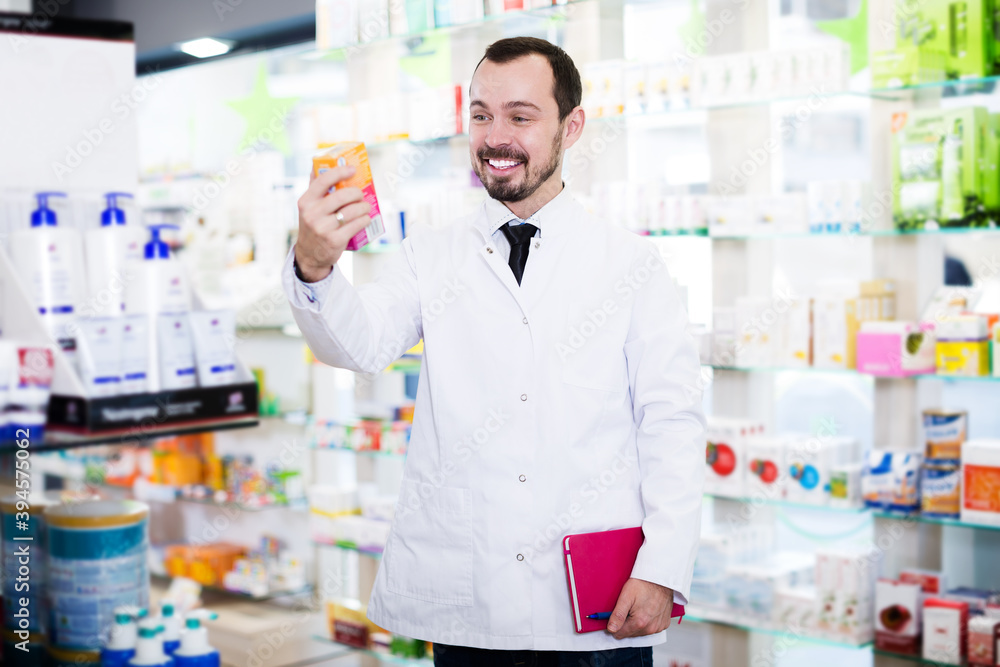 Young positive male pharmacist noting assortment of drugs in pharmacy