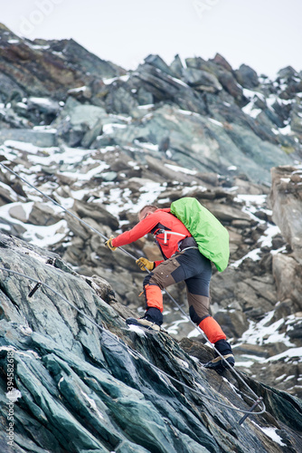 Brave alpinist holding rope while climbing alpine ridge. Young climber with backpack ascending mountain and trying to reach mountaintop. Concept of mountaineering, alpinism and alpine climbing.