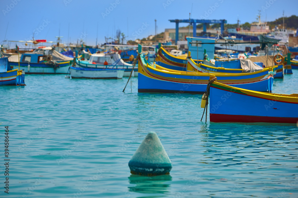 Obraz premium The colorful Blue-yellow fishing boats on the turquoise sea against orange ancient buildings. Vivid colors. Fishing port, view from the water surface. Island of Malta. 