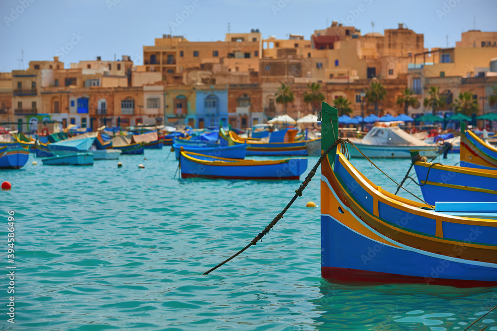  Blue-yellow fishing boats on the turquoise sea against orange ancient buildings.  Malta.
