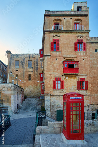A deep red telephone kiosk on the street of the ancient city of Valletta in evening colors. Vertical, no people. Postcard. Holidays in Malta. © Martin Mecnarowski