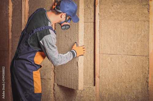 A man in overalls puts mineral sheet insulation between wooden beams. The concept of insulation or sound insulation of walls in frame buildings.