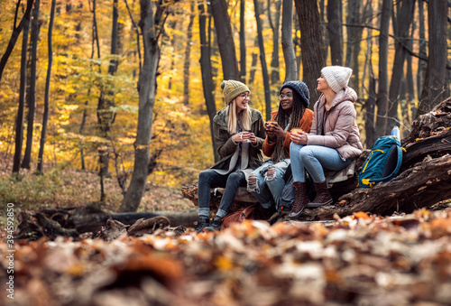 Three female friends resting after hiking in forest sitting on collapsed trunk and drinking tea.