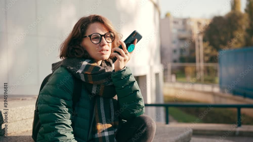 A young woman in glasses recording a voice message on her phone while sits on the stairs of a building on the street on a sunny autumn day. A young woman in glasses recording a voice message on her phone while sits on the stairs of a building on the street on a sunny autumn day.