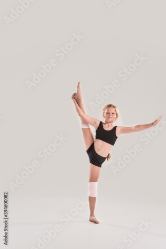 Full length shot of cute little redhead girl gymnast looking at camera, raising one leg with her hand, showing flexibility isolated over grey background