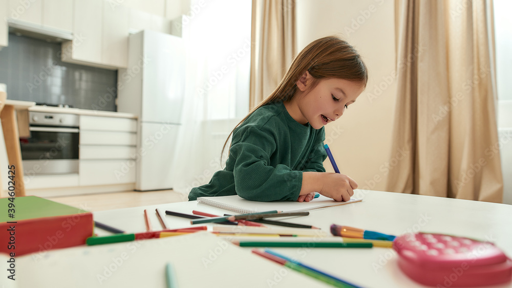 A cute small girl enjoys speaking and writing with a pencil while sitting at a table in a big bright kitchen A cute small girl enjoys speaking and writing with a pencil while sitting at a table in a big bright kitchen