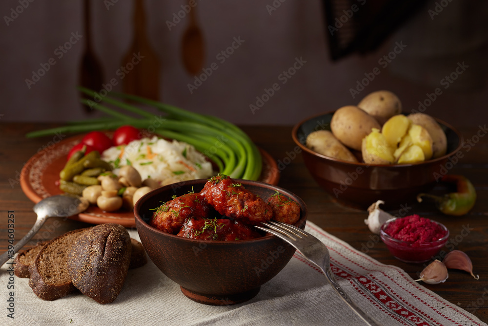Meat balls in tomato sauce in a ceramic plate on a wooden table with potatoes and snacks. Rustic dinner with meatballs and snacks Meat balls in tomato sauce in a ceramic plate on a wooden table with potatoes and snacks. Rustic dinner with meatballs and snacks