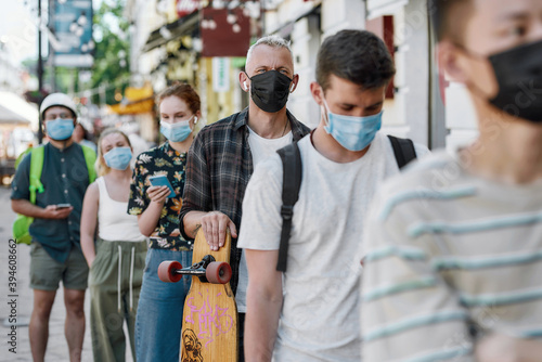 Middle aged man holding longboard, wearing mask waiting, standing in line, respecting social distancing to collect his takeaway order from the pickup point during lockdown photo