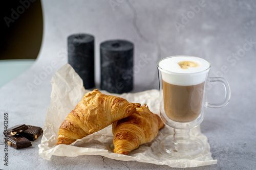 On a gray table there is a glass cup with hot latte coffee and next to it two croissants three pieces of chocolate and black candles