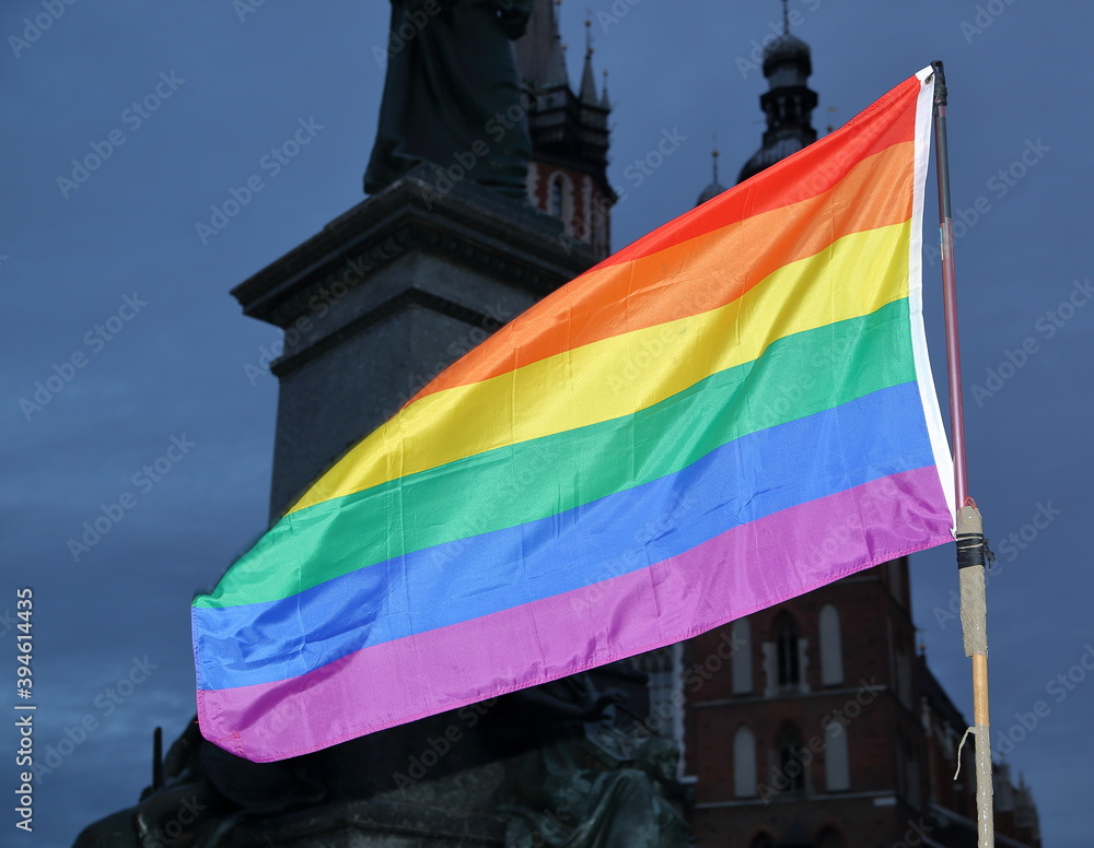 flag of rainbow symbol LGBT+ groups waves on pole against Adam Mickiewicz monument and Saint Marys church in Krakow, Poland