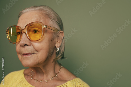 Close up portrait of senior thoughtful serious woman with gray hair, big stylish glasses, earings and pearls. Neutral old woman wearing yellow shirt looking away. Isolated over green background.