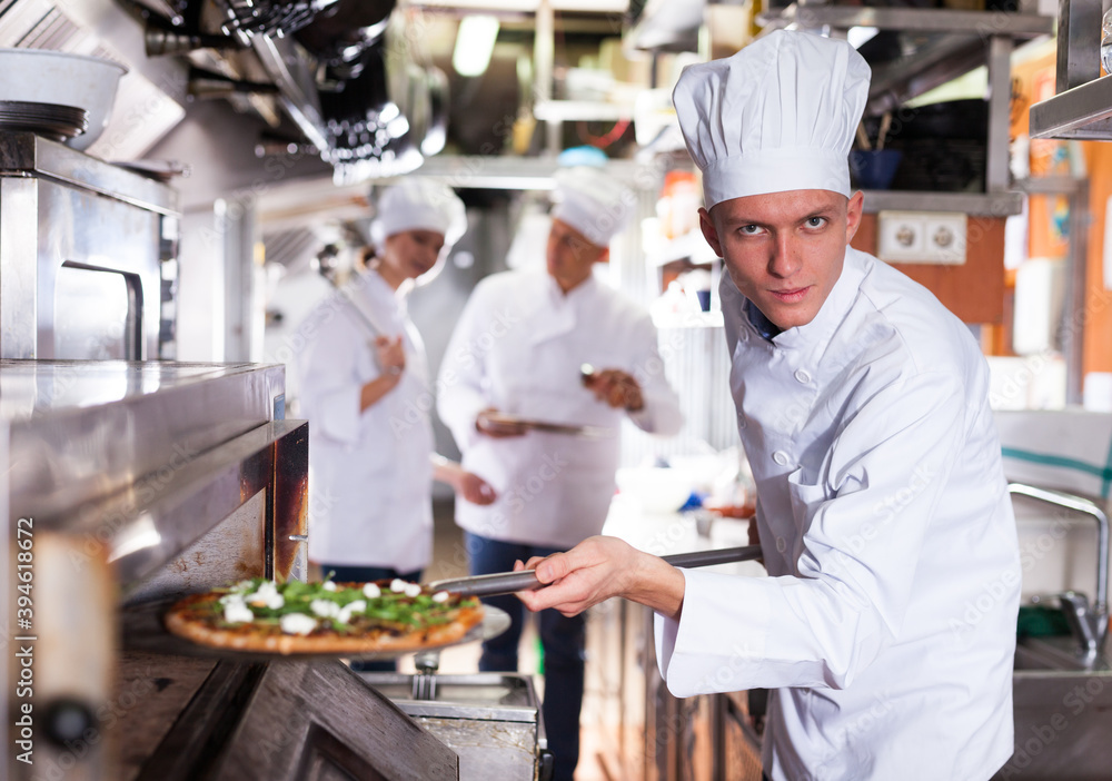Young male chef in white uniform working in professional kitchen of restaurant, baking ordered pizza