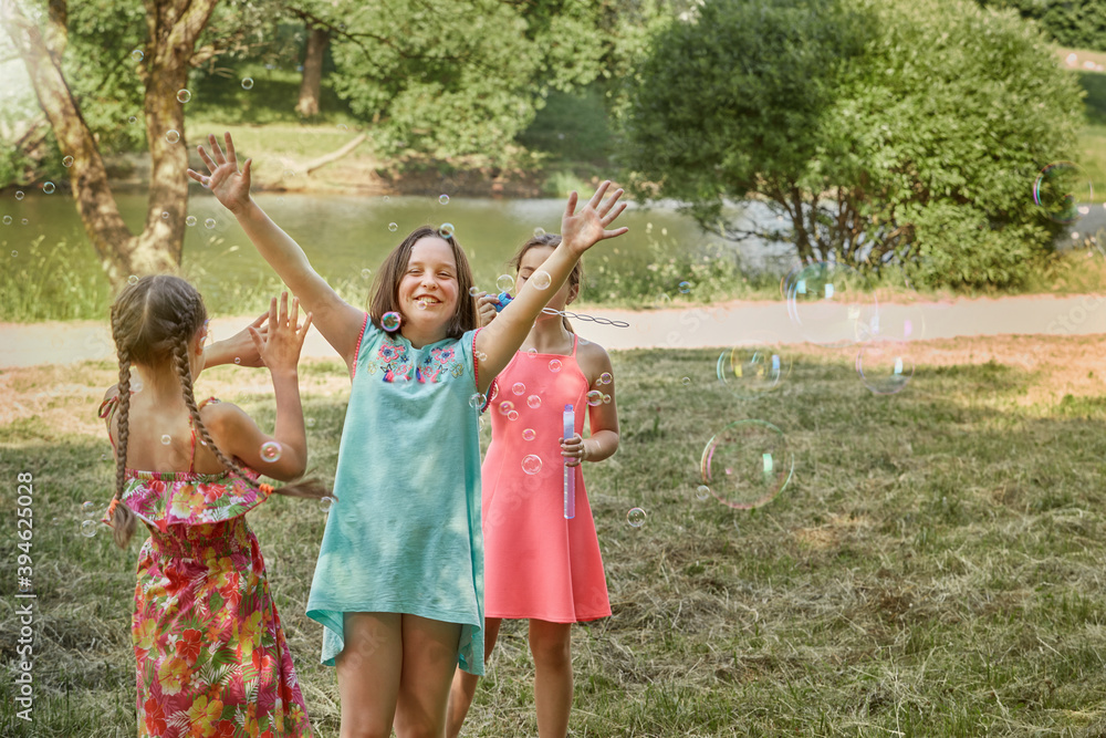 Fototapeta premium Beautiful girls play together in the Park and blow soap bubbles. On the background of a large lake.