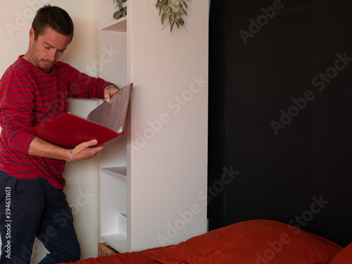 MAN IN RED T-SHIRT, BLUE TROUSERS AND BLACK GLASSES, IN FRONT OF THE WHITE SHELF OF HIS ROOM, LOOKING AND TAKING A NOTEBOOK