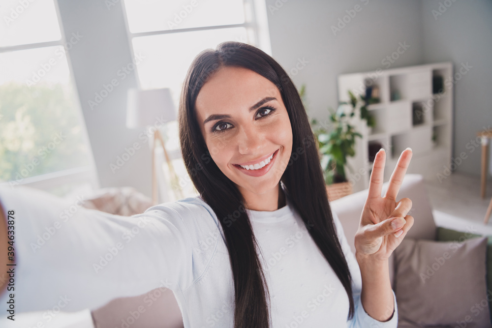 Photo of happy joyful attractive sweet young woman make camera selfie in living room show v-sign indoors in house Photo of happy joyful attractive sweet young woman make camera selfie in living room show v-sign indoors in house