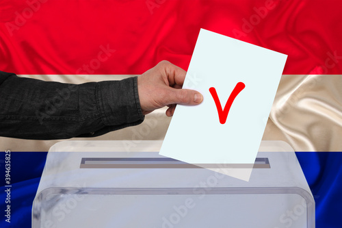 male voter drops a ballot in a transparent ballot box against the background of the national flag of the Netherlands, concept of state elections, referendum