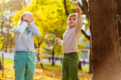Cute blond little boy and older girl enjoying an autumn day in nature, kids playing together with big flying soap bubbles. Adorable brother and sister trying to catch and burst a big bubble.
