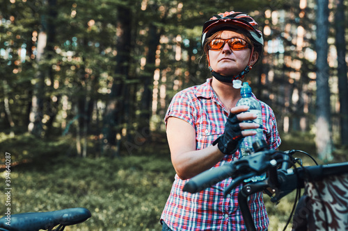 Active woman spending free summer vacation time on a bicycle trip in a forest. Woman wearing bicycle helmet and gloves holding bike with basket and drinking a water from bottle #394640287