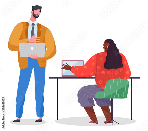Man in strict suit stands and holds laptop, dark-skinned woman at table with laptop. Office workers at business meeting, discussing project. Coworkers work together. Mutually beneficial cooperation