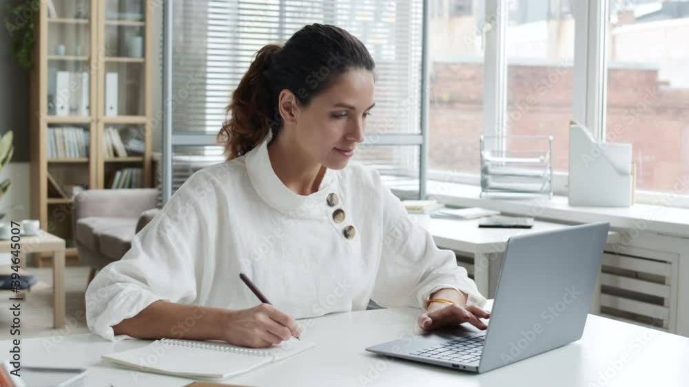 Lockdown of young Caucasian woman with curly hair sitting at workplace, working on laptop and making notes in notebook Lockdown of young Caucasian woman with curly hair sitting at workplace, working on laptop and making notes in notebook
