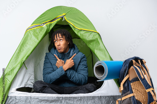 Young african american man inside a camping green tent pointing to the laterals having doubts