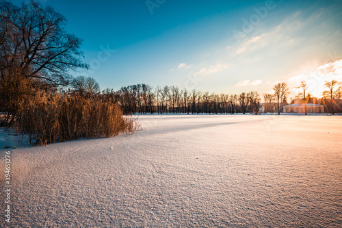 Winter landscape. Scenic view over frozen lake at sunset. Leafless trees and bushes, reed grass and icy ground surface. Pavilion building on the shore. Sunny day city in park. Cold weather. Copy space