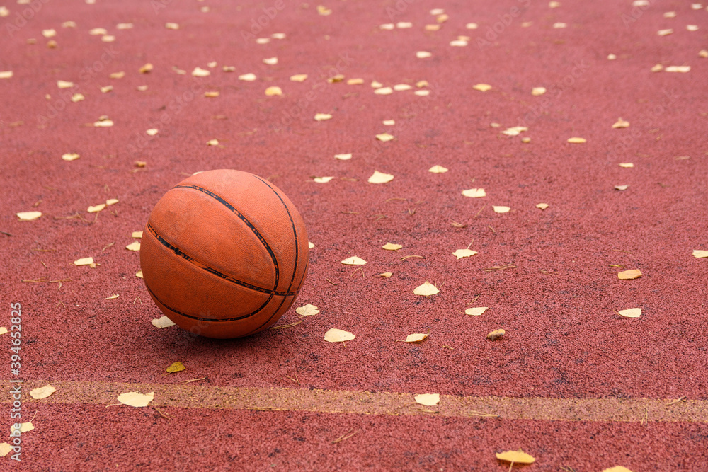 An old battered basketball is lying on the outdoor court at the autumn day. Plenty of fallen leaves on the court. Low angle view An old battered basketball is lying on the outdoor court at the autumn day. Plenty of fallen leaves on the court. Low angle view