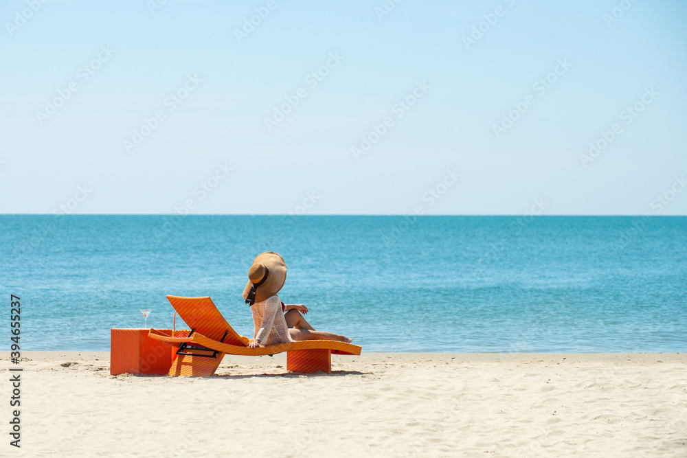 Happy young woman in bikini and sun hat resting on beach chair for sunbathing and looking beautiful nature of the sea on island beach in sunny day. Pretty girl relax and enjoy summer holiday vacation.