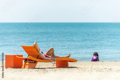 Happy Asian family holiday vacation. Asian mother resting on beach chair looking little daughter playing on the beach in summer day. Cute child girl relax and having fun together with mom on the beach