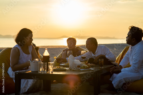Group of multi generation Asian family enjoy dinner party together on the beach at sunset. Diverse family with child girl, adult and senior couple relax and having fun on summer holiday vacation
