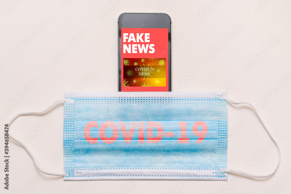 Fake news concept on coronavirus theme. Mobile phone and medical mask with COVID-19 text. News feed on smartphone screen with fake articles Fake news concept on coronavirus theme. Mobile phone and medical mask with COVID-19 text. News feed on smartphone screen with fake articles