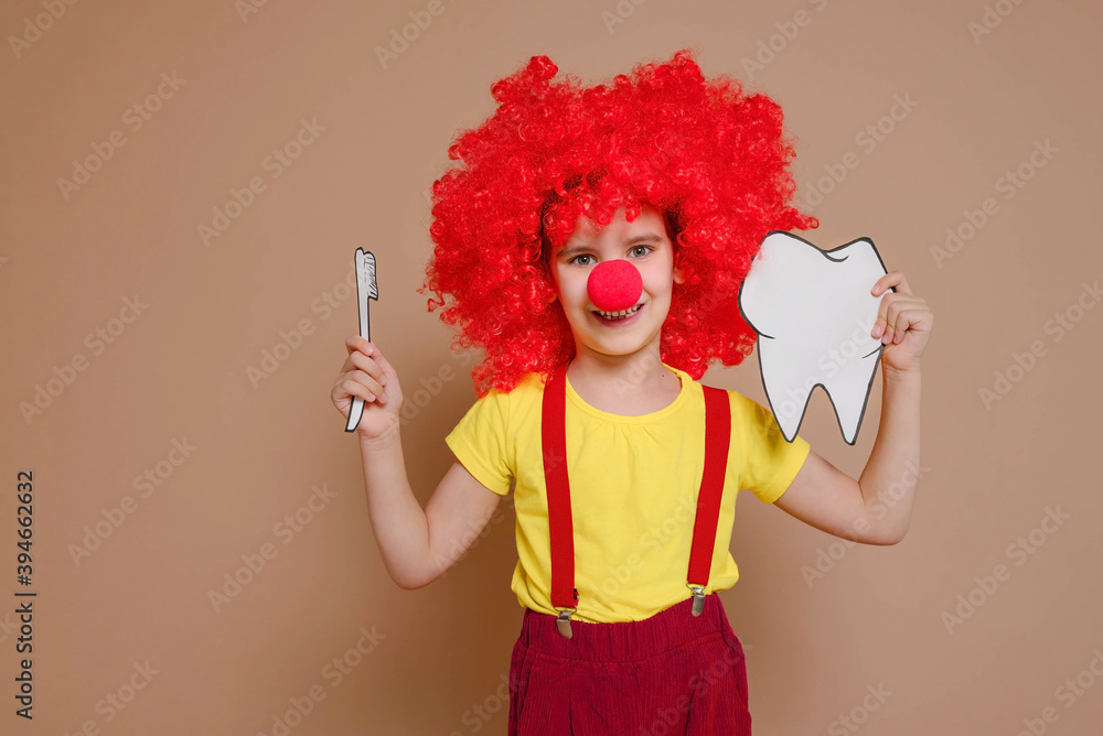 Funny clown girl child in a red wig and with a nose holding a mock-up of a tooth and brush isolated against the background of a Set Sail Champagne flower. Kid teeth hygiene, teeth cleaning concept. Funny clown girl child in a red wig and with a nose holding a mock-up of a tooth and brush isolated against the background of a Set Sail Champagne flower. Kid teeth hygiene, teeth cleaning concept.