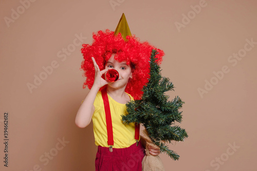 Funny clown girl in a red wig and with a nose made of a Christmas ball-toy holds a Christmas tree isolated against the background of a Set Sail Champagne flower. Holidays, circus concept.