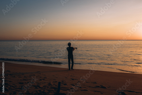 Silhouette of a man fishing on a background of beautiful colorful sunset next to the pleasant sea on the sandy beach.