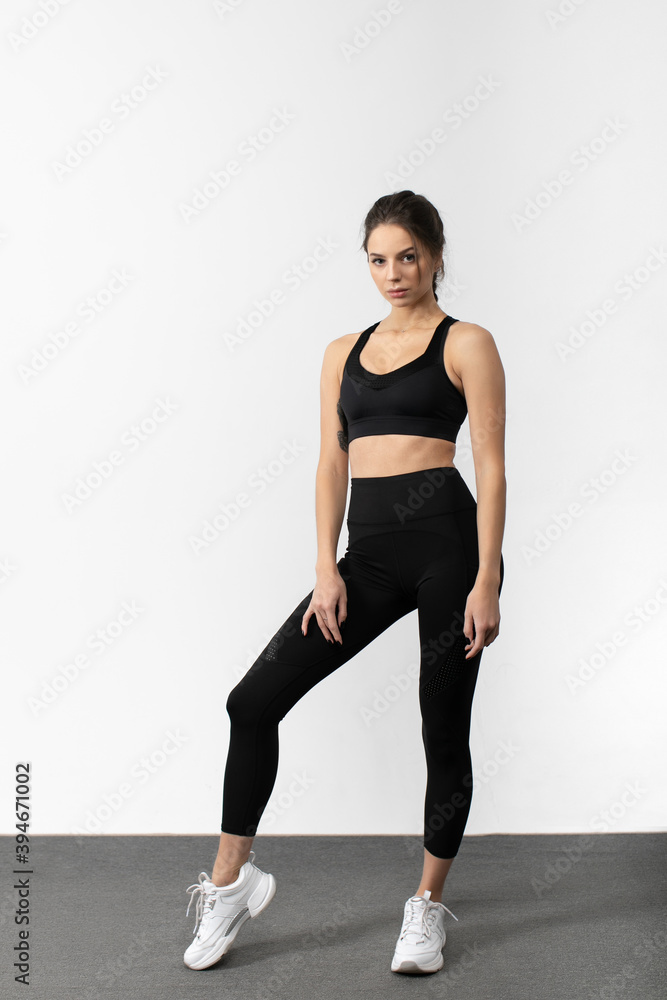 Sporty slim woman in sportswear stands in gym and poses against white background