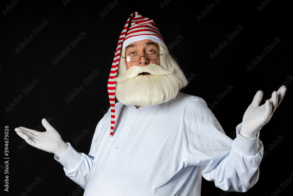 A bearded man Santa Claus in a nightgown and glasses looks with delight, spreading his arms from the side. On a black background.