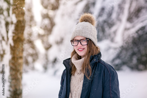A young woman with glasses, a knitted hat with a pompom and a warm sweater , stands in the winter forest and smiles happily