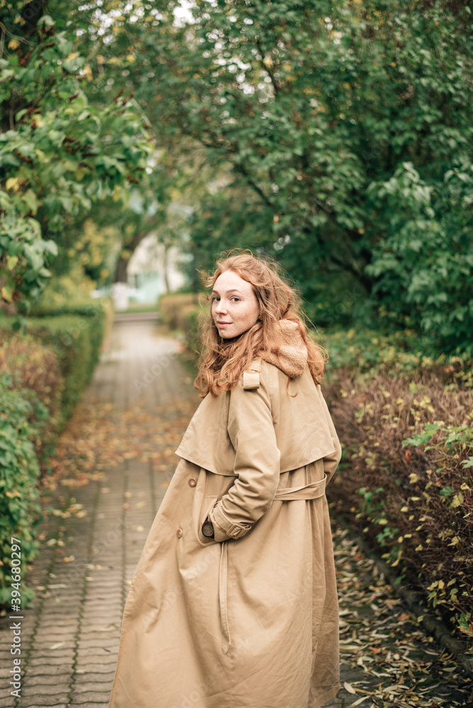 Ginger girl in a trench coat walks through the park and looks back at the camera Ginger girl in a trench coat walks through the park and looks back at the camera
