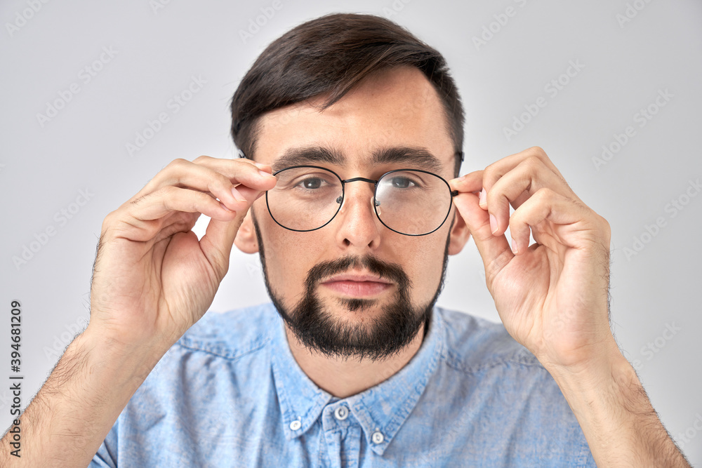 Studio shot caucasian bearded man with poor eyesight holds hands on the rim of glasses and squints, trying to see something, looking at the camera isolated on blue background Studio shot caucasian bearded man with poor eyesight holds hands on the rim of glasses and squints, trying to see something, looking at the camera isolated on blue background