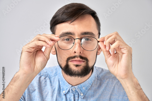 Studio shot caucasian bearded man with poor eyesight holds hands on the rim of glasses and squints, trying to see something, looking at the camera isolated on blue background