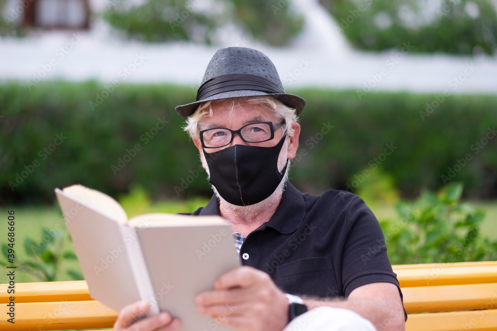 Portrait of smiling senior man wearing black face mask due to coronavirus sitting on a yellow bench in public park reading a book. Old people with black hat looking at camera