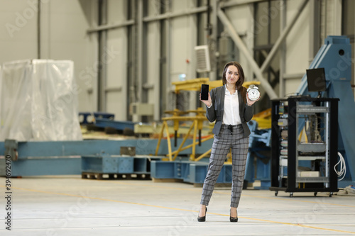 Portrait of a female factory manager in a business suit holding mobile phone and retro alarm clock , controlling the work process in the helicopter manufacturer. Timeline. Deadline. 