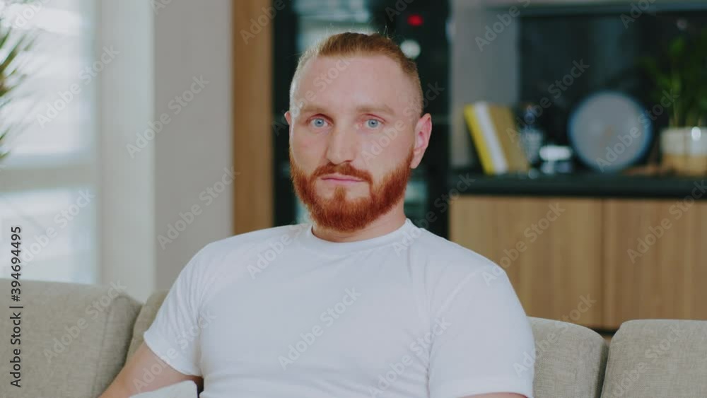 Close up serious man sitting on couch at home. Looking at the camera focused in modern sunlight apartment. Confident comfortable cozy. Slow motion Close up serious man sitting on couch at home. Looking at the camera focused in modern sunlight apartment. Confident comfortable cozy. Slow motion
